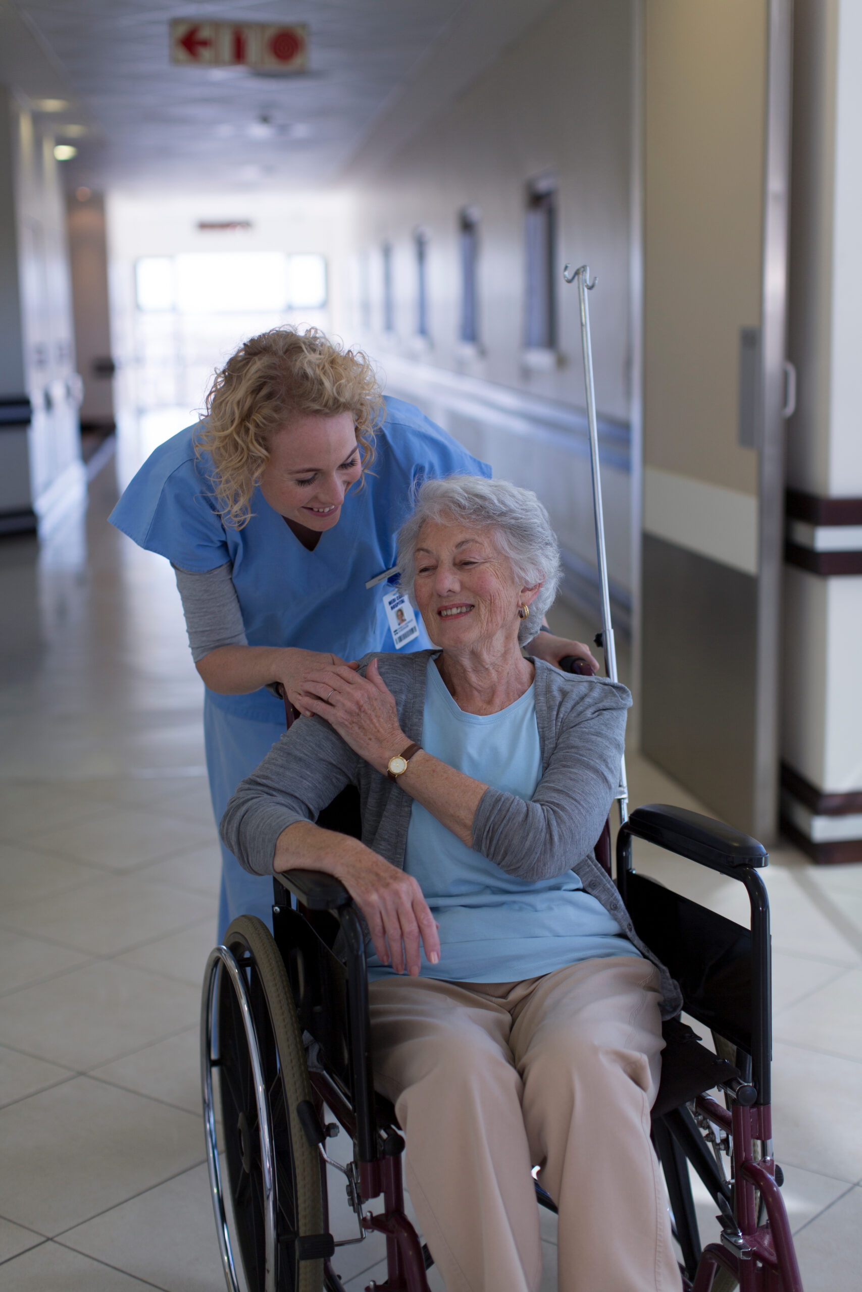 Female nurse comforting senior patient in wheelchair in hospital corridor